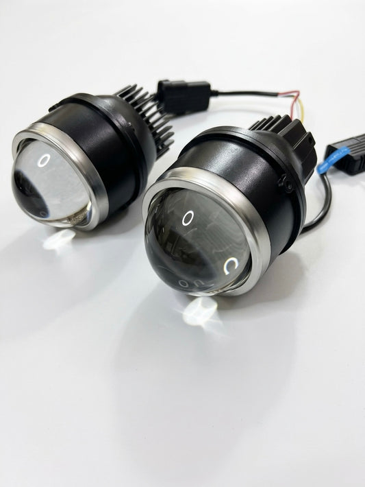 Two black automotive LED headlight pods with clear lenses and silver trim, shown on a white reflective surface with their wiring harnesses visible.