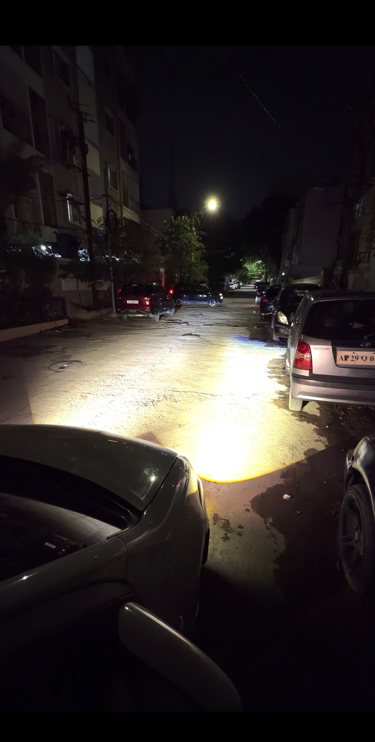 A car's bright front headlight illuminating a wet residential street at night, with parked cars and apartment buildings visible in the dark surroundings.