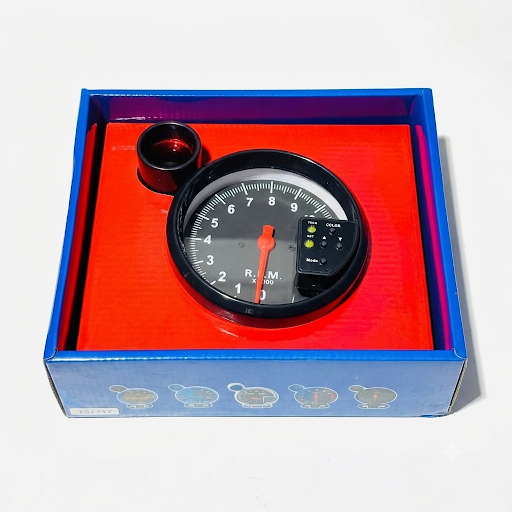 A car gauge kit with a black analog tachometer-style gauge, red needle, and small black lens piece, displayed inside an open blue box with a bright red insert on a white background.